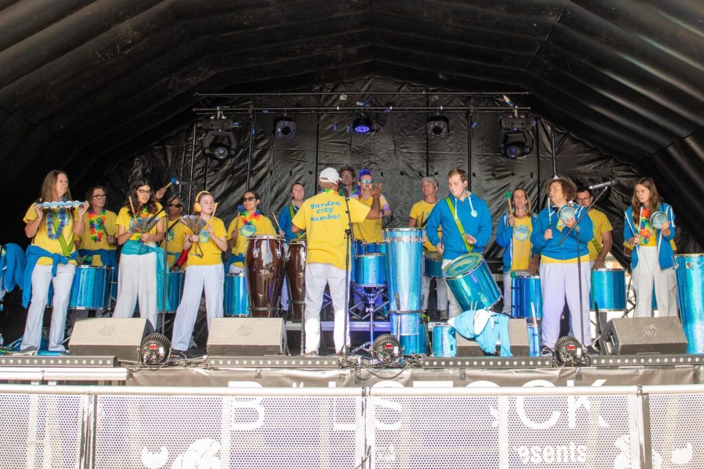 GCS drummers performing a gig on a tall outdoor stage with a rounded arch cover. Band members wearing yellow t-shirts and white trousers, playing various instruments.