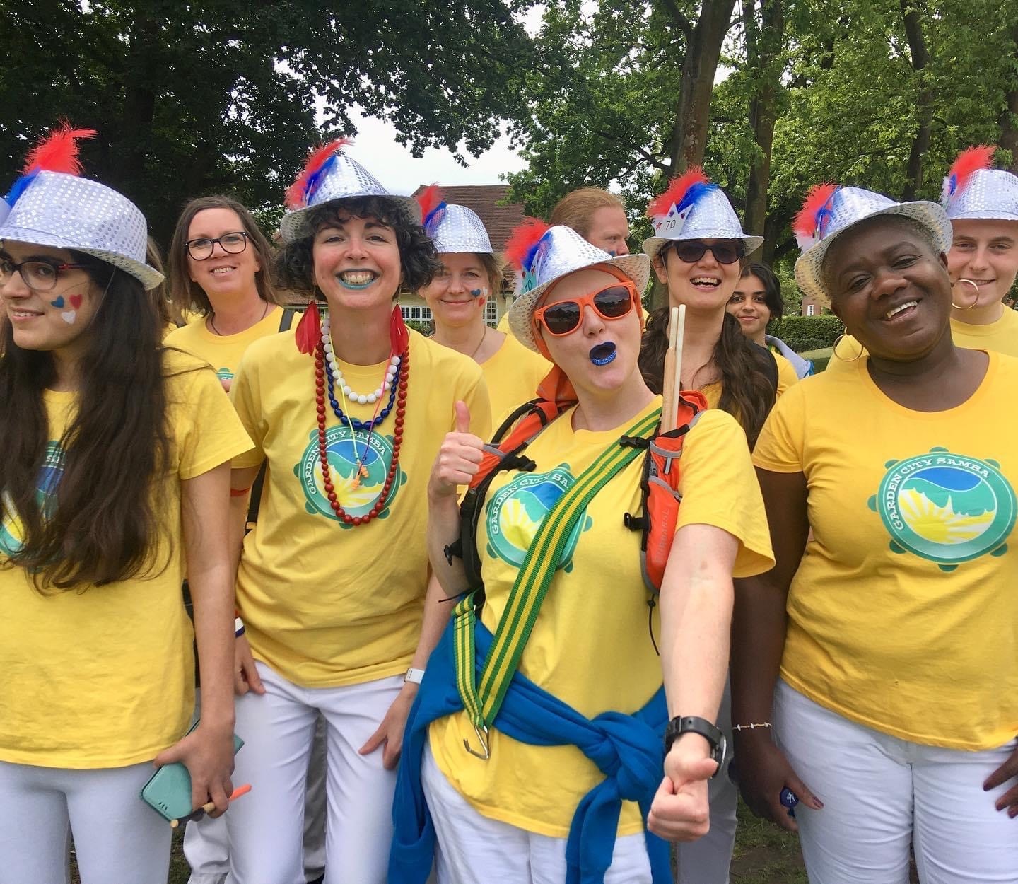 Smiling group photo of GCS before a gig. Wearing yellow t-shirts and white trousers, plus silver sequined hats with red and blue feathers attached.