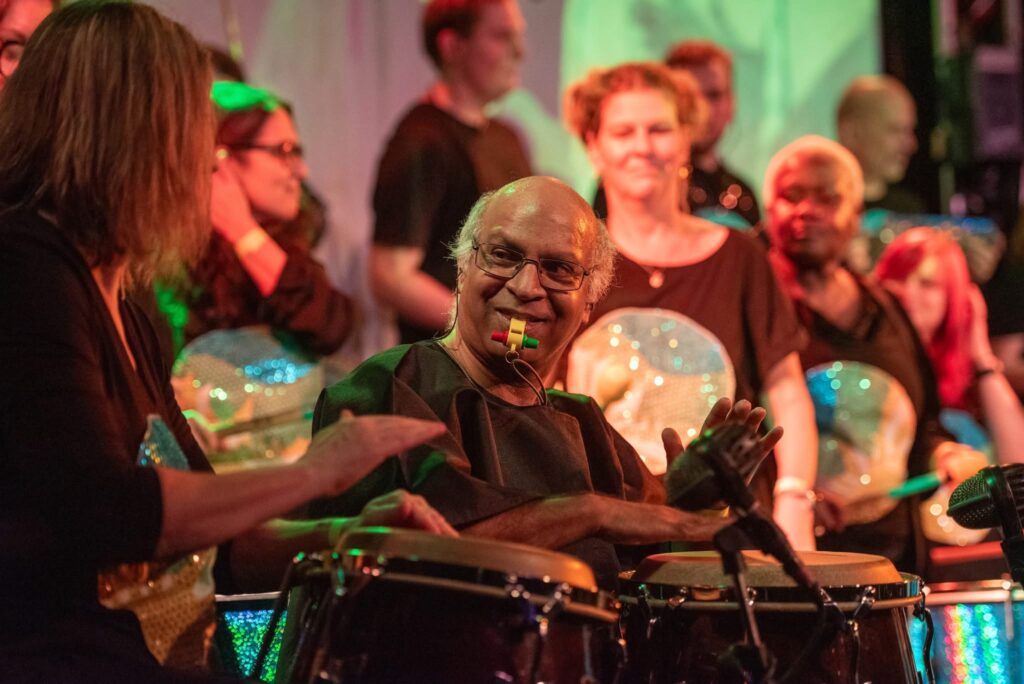 Ravin Jayasuriya, a South Asian man, smiles while playing congas on an indoor stage with GCS members behind him holding instruments. All performers are wearing black with sequined GCS logos on the front.