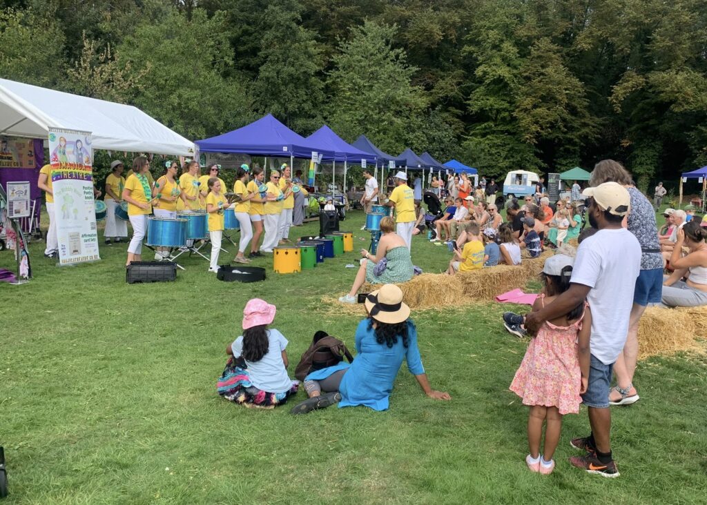 GCS drummers perform at a summer fate type event in a field while families watch on. Performers play a variety of instruments and wear yellow t-shirts with white trousers