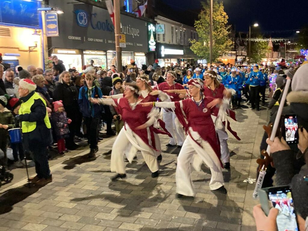 GCS parade through a busy town center lit by street lamps and led by six dancers in maroon and white costumes. Drummers wear black trousers with blue hoodies and silver sequinned hats adorned with multi-coloured lights. They play a variety of instruments.