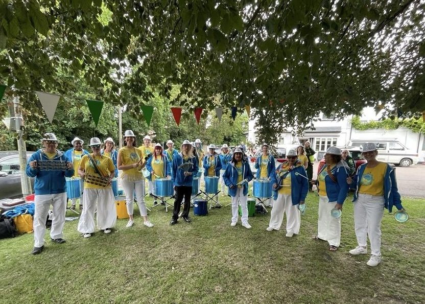 GCS drummers smile posing for a photo with their instruments, under a tree. They are wearing white trousers, yellow t-shirts and silver sequined hats.