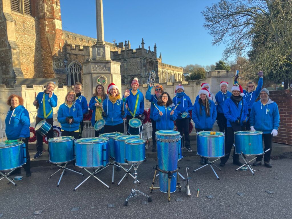 GCS drummers smile posing for a photo with their instruments. They wear black trousers and blue hoodies and some wear santa hats.