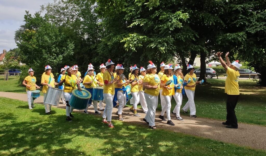 GCS drummers parade through a sunny park wearing yellow t-shirts, white trousers, and silver sequined hats with blue and red feathers attached. They are playing a variety of instruments. A man directs the group from the front facing towards them, his arms raised mid-signal.