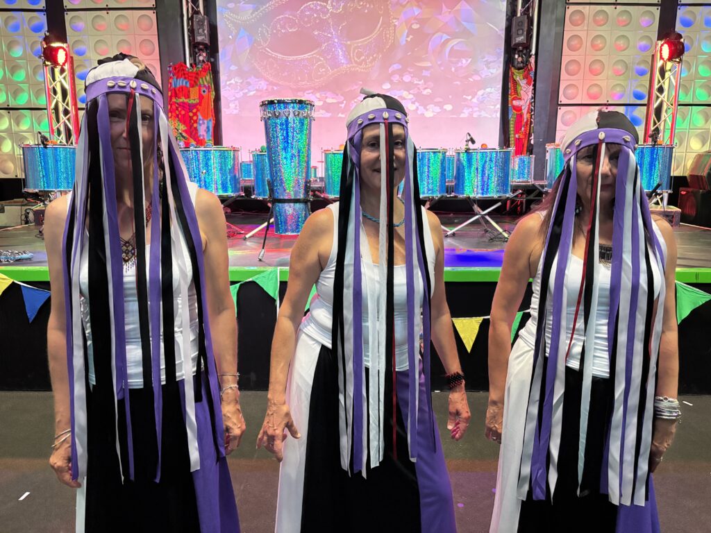 Three dancers smile for a photo in front of an indoor stage. They are wearing black, white, and purple costumes with long stranded headdresses that partially obscure their faces.