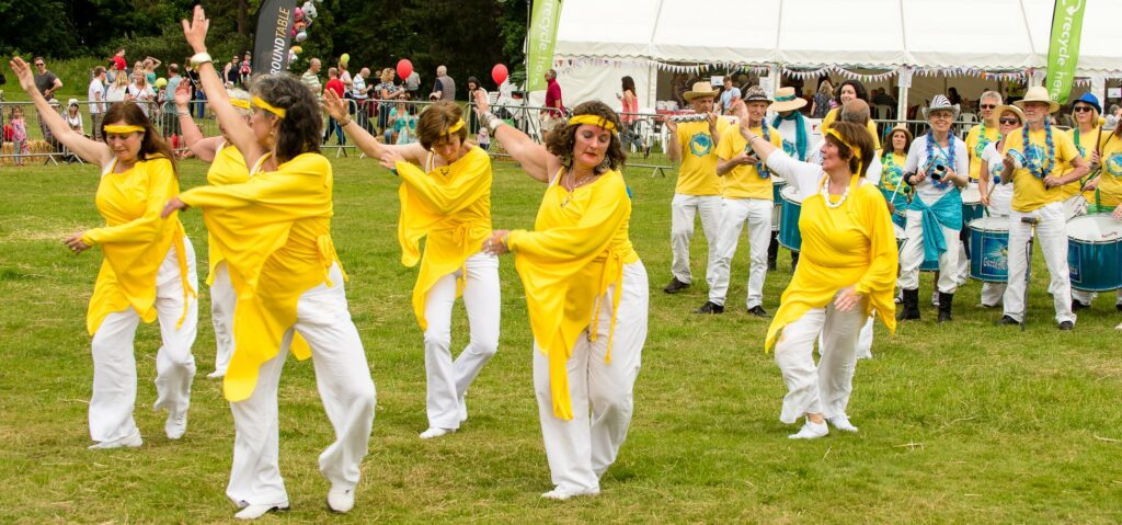 A group of dancers in yellow and white costumes dance in a field ahead of a samba band.