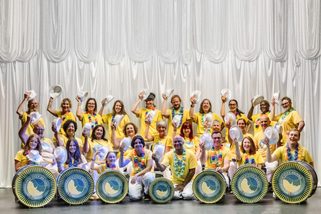 Professional group photo of the band smiling in front of a silvery curtain backdrop. They are wearing yellow t-shirts, white trousers, multicoloured garlands, and silver sequined hats held to one side as if in greeting. Some drums are placed in front.
