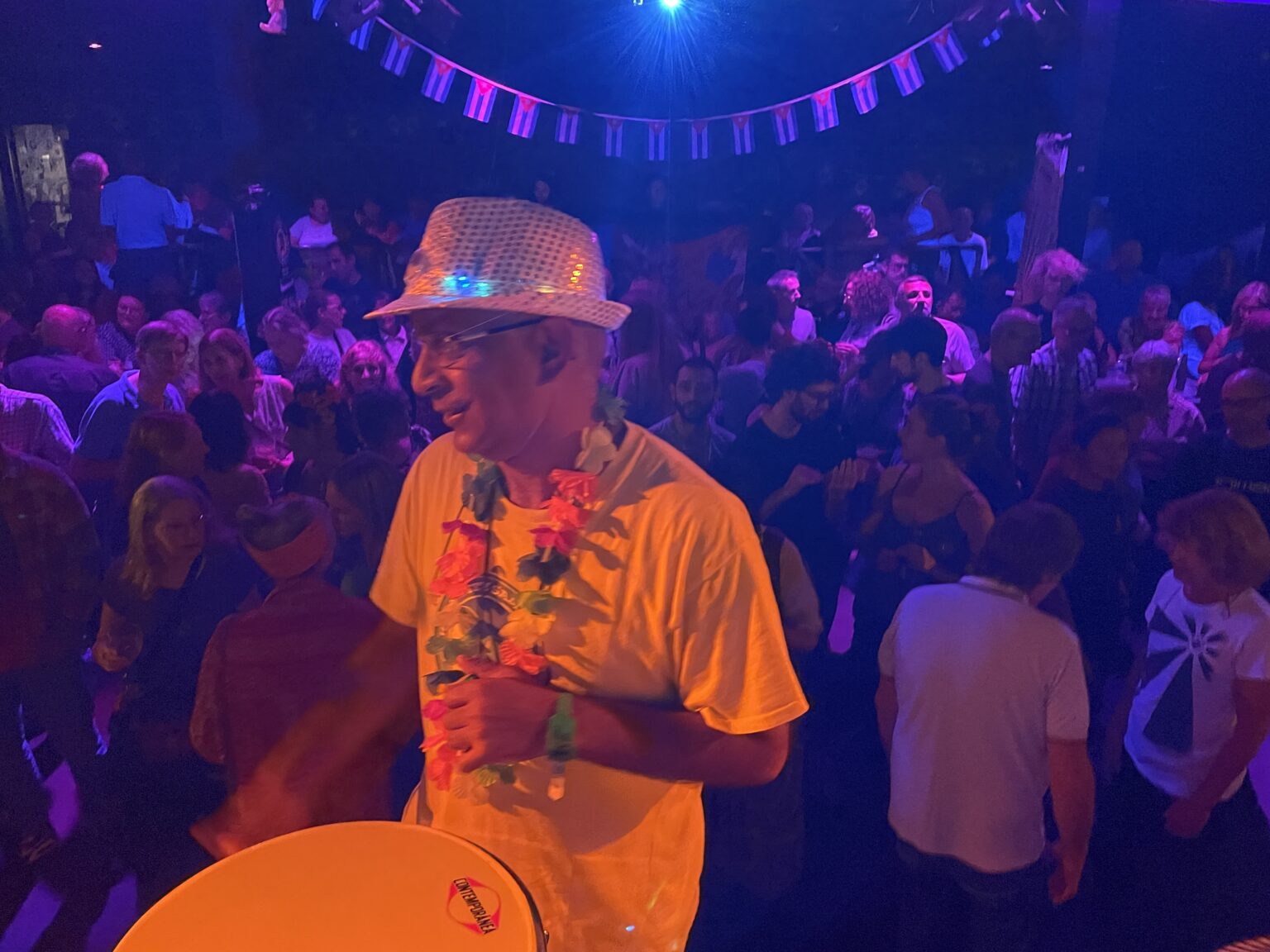 Smiling band leader from the band's perspective, drumming on an indoor stage with a packed crowd in the background. Wearing a yellow t-shirt, white trousers, multi-coloured garland and sliver sequined hat with lights.