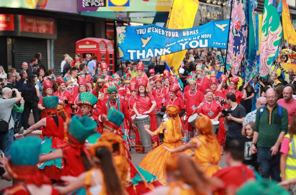 A sea of drummers and dancers parade through crowded a sunny street. Performers are wearing brightly coloured costumes in a mix of red, blue, and yellow.