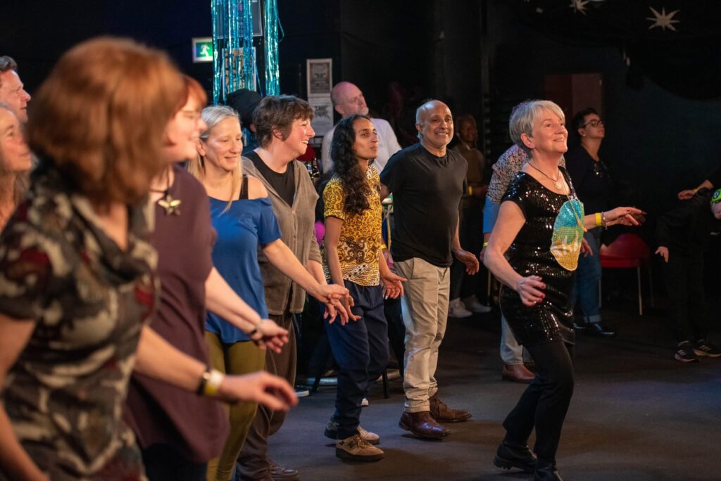 A group of people copy choreography from a woman dancing in front wearing a black shirt with a GCS logo on.