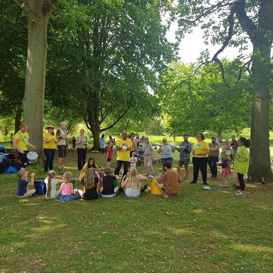 Children sit in a circle in a tree-filled park playing various instruments and watching GCS band members for instructions