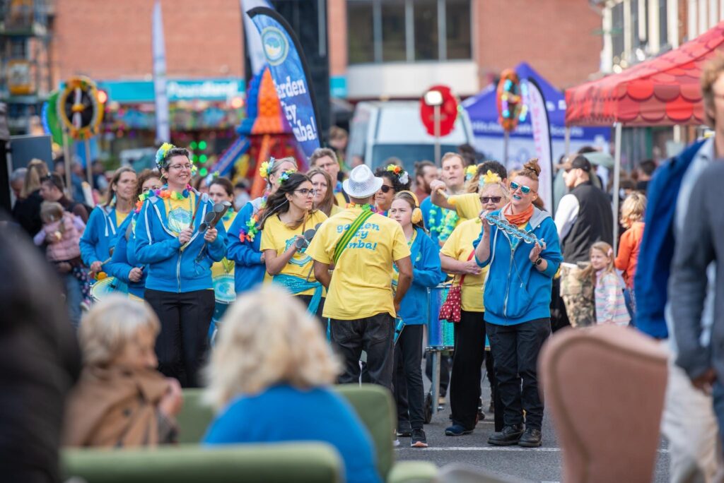 GCS drummers happily parading through a crowded street playing various instruments. Performers wear yellow t-shirts, black trousers and flowers pinned in hair.