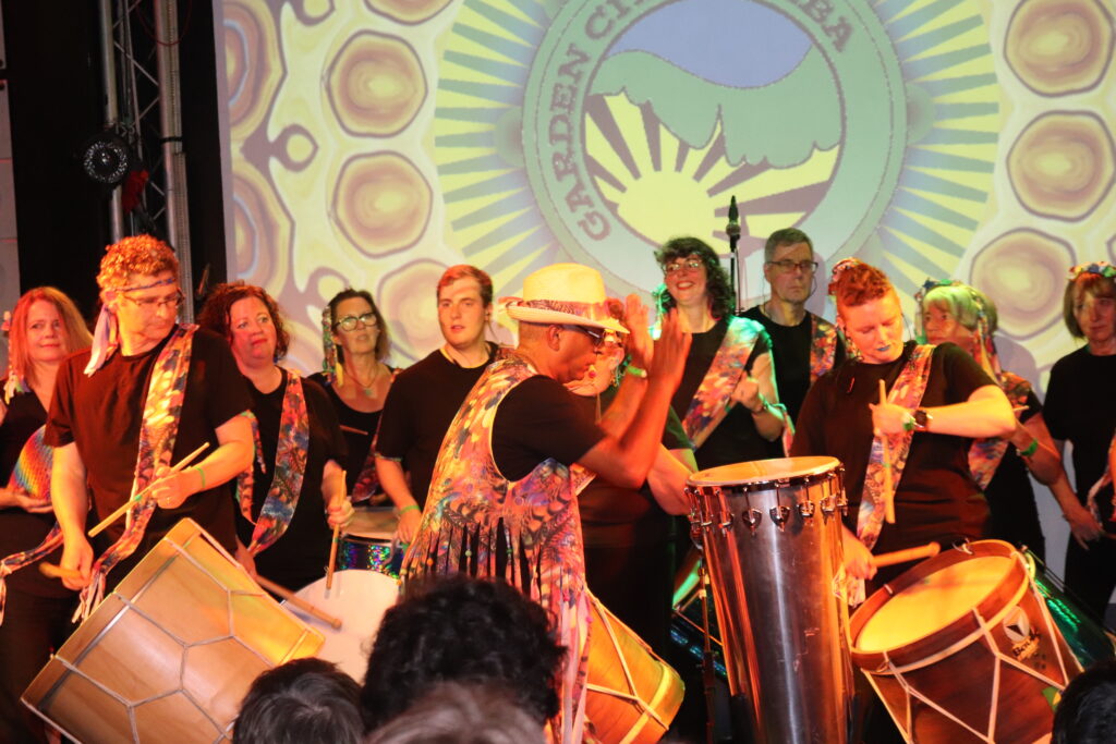 GCS performing on a stage in a club dressed in black with colourful sashes. Band leader is wearing a colourful waistcoat in with lots of beaded tassels.
