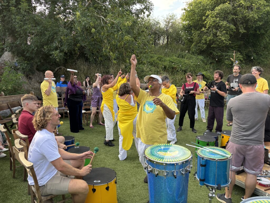 GCS drummers and dancers lead a workshop for an audience in a clearing surrounded with trees. The audience are clearly enjoying getting to play a variety of instruments.