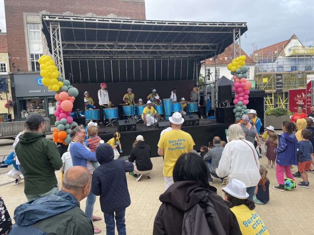 Families gather on and around an outdoor stage to take part in a samba drumming workshop led by various GCS members in yellow t-shirts and white trousers.