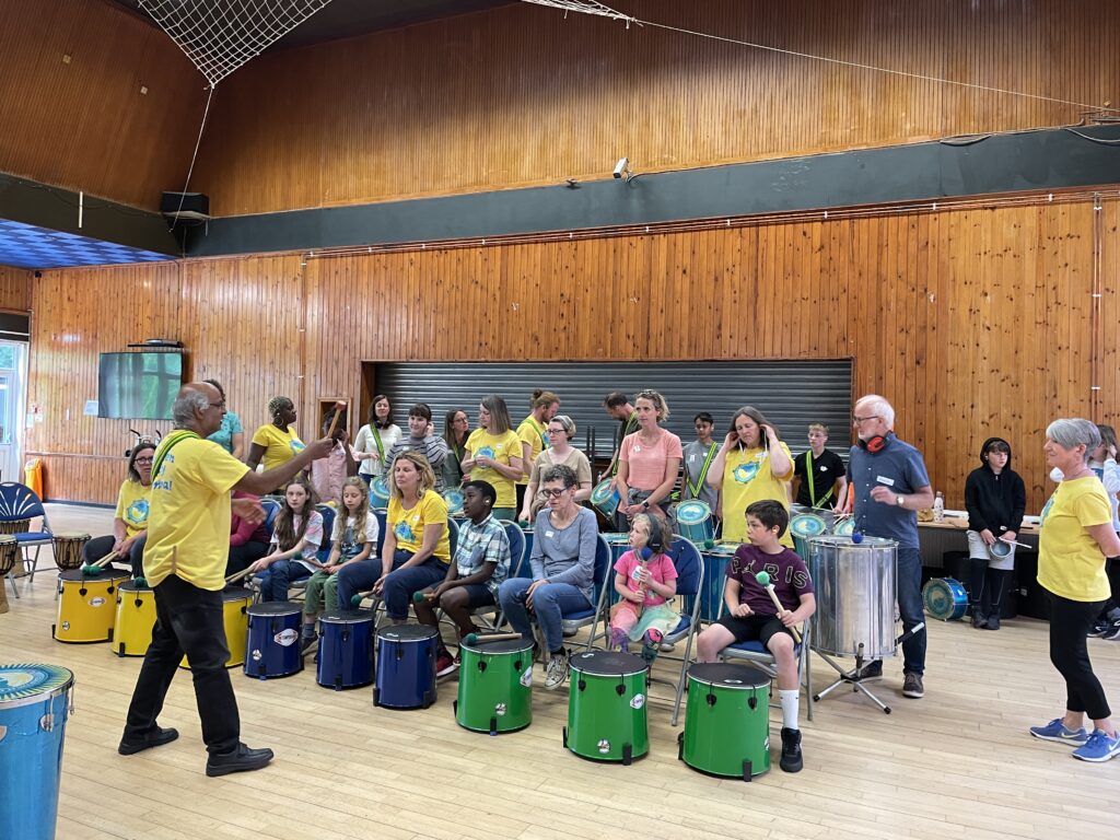 A group of people of all ages is led in a samba drumming workshop in a community hall. Various members of GCS are present wearing yellow t-shirts.