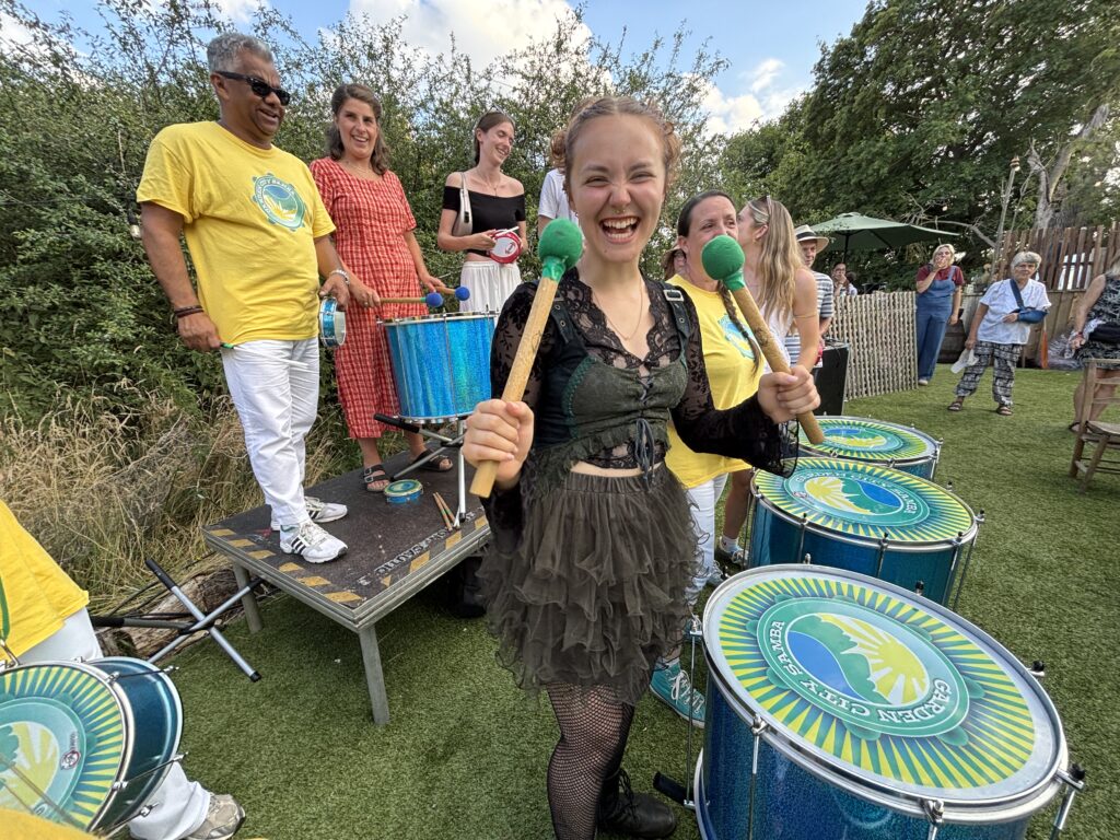 A smiling member of the public waves drum beaters towards the camera during a samba drumming workshop led by various GCS members in yellow t-shirts and white trousers.