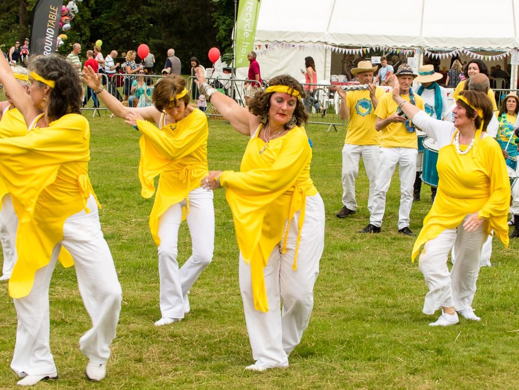 A group of dancers in yellow and white costumes dance in a field ahead of a samba band.